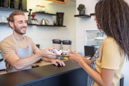 Glass of coffee. Cheerful man in apron behind bar giving glasses of take-away coffee to woman in cafeの写真素材