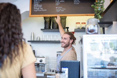 Cafe, coffee selection. Joyful young bearded man in apron pointing at menu with variety of coffee hanging on wall and womans backの写真素材