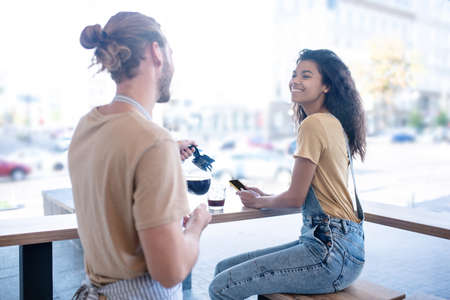 Time for coffee. Happy woman in denim overalls sitting in cafe near large window and man in apron with jug of coffeeの写真素材