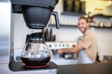 Coffee maker. Coffee maker with glass jug of coffee standing on counter and man at distance in coffee shopの写真素材