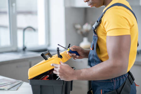 Home repair. Dark-skinned service man standing in the kitchen and holding a box with tools
