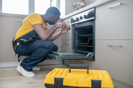 Gas stove repair. Dark-skinned service man in yellow tshirt repairing the gas stoveの写真素材