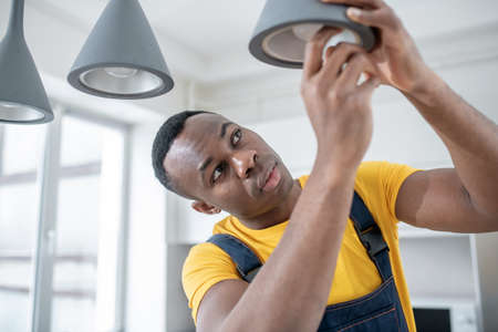 Electrician. Dark-skinned service man in yellow tshirt repairing electricity lampsの写真素材