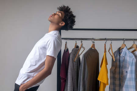 Curly-haired young guy standing near the hangers with clothes and looking thoughtfulの写真素材