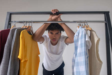 Curly-haired young guy standing near the hangers with clothes and looking thoughtfulの写真素材