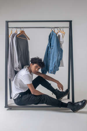Curly-haired young guy sitting near the hangers with clothes and looking tiredの写真素材