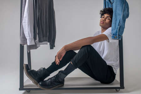 Curly-haired young guy sitting near the hangers with clothes and looking tired
