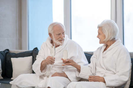 Senior couple in white robes drinking champagne and looking pleasedの写真素材