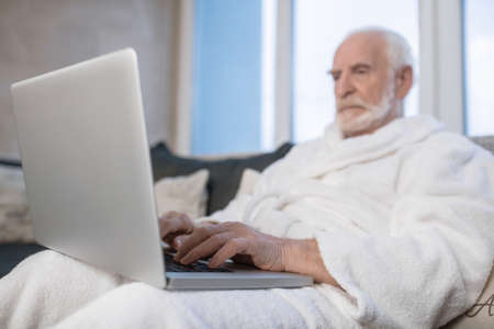 Gray-haired bearded man in white robe working on laptop and looking seriousの写真素材