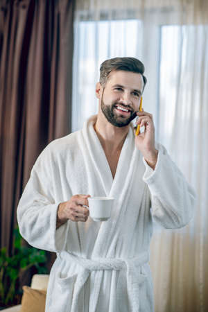 Young bearded man in a white bath robe having morning coffee and talking on the phone