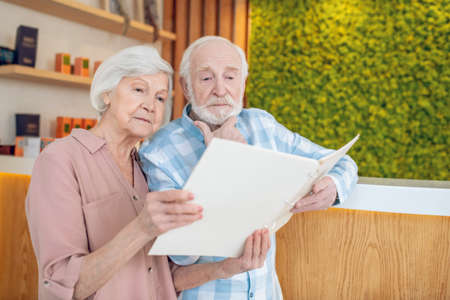 Gray-haired couple standing near reception in a spa center and looking trhough the price listの写真素材