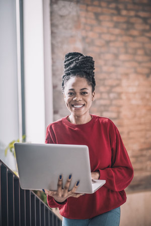 Black woman in red shirt with a laptop smiling and walking in the officeの写真素材
