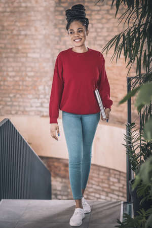 Black woman in red shirt with a laptop smiling and walking in the officeの写真素材