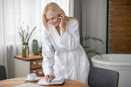 Long-haired woman in a white bath robe talking on the phone and making notesの写真素材