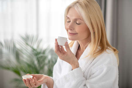 A blonde woman in a white bath robe holding a jar of a new cream
