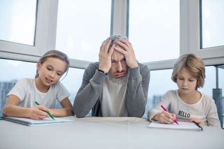 Gray-haired bearded man sitting with his kids at the table while they doing lessons
