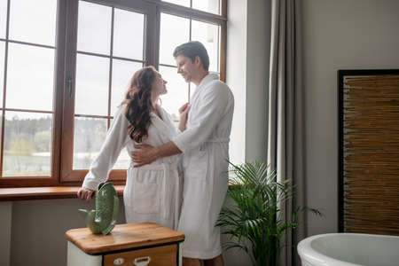 Two people in white bathrobes standing near the window and looking affectionateの写真素材