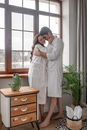 Two people in white bathrobes standing near the window and looking affectionateの写真素材