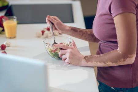 A dark-skinned woman eating salad and watching something on laptopの写真素材