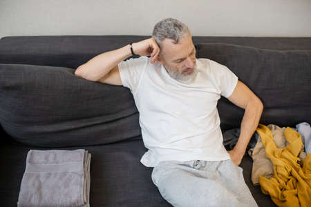 A gray-haired man sitting on the sofa and looking at the clothes lying near