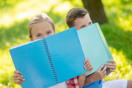 Girl and boy in playful mood hiding behind notebooksの写真素材