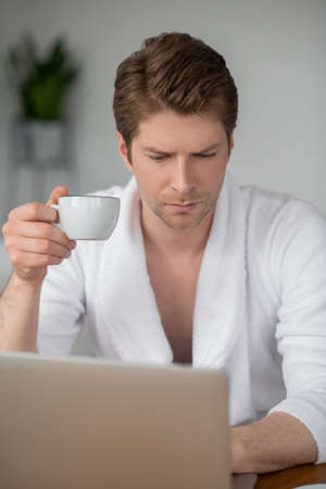 A handsome man in a white bathrobe sitting at the laptop and spending time onlineの写真素材