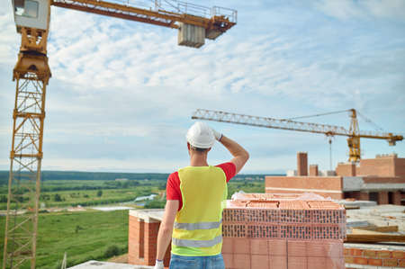 Man in a hardhat standing before cranes and stacked red bricksの写真素材