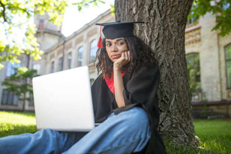 A girl in academic gown sitting under the tree with a laptopの写真素材