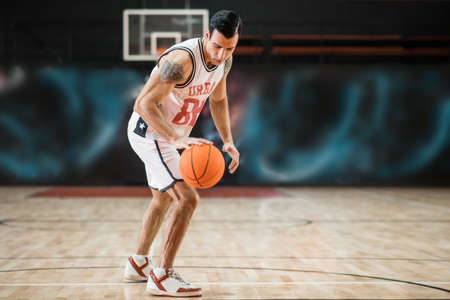 Young athletic man in white sportwear playing basket-ball in the gymの写真素材