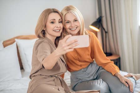 Two smiling blonde women sitting on the bed and making selfie on a smartphone