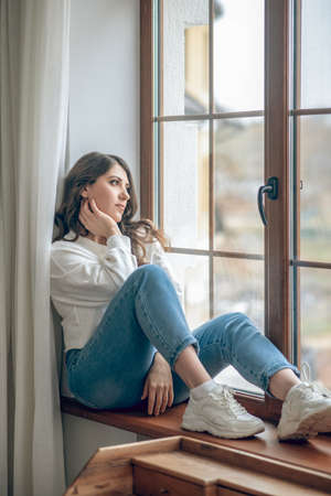 Pretty young long-haired woman in a white blouse sitting at the window and looking thoughtfulの写真素材