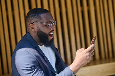 A dark-skinned man in a suit sitting on the bench with a smartphone in handsの写真素材