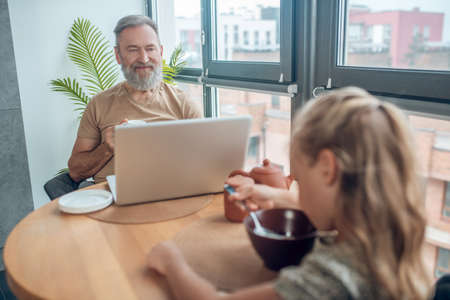 A cute blone girl eating her breakfast while her dad working on laptopの写真素材