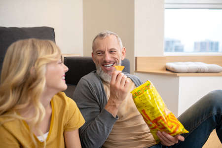 A blonde teen sitting on the floor with her dad and eating chips