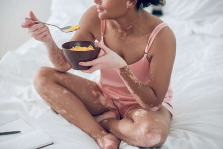 A dark-skinned woman eating cereals while sitting on bedの写真素材