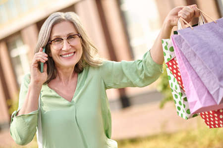 Woman with smartphone near ear showing shopping bagsの写真素材