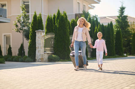 Blonde woman carrying a suitcase and holding her daughters handの写真素材
