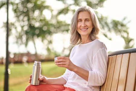 Woman with thermos sitting on bench looking at cameraの写真素材