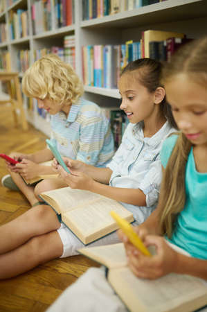 Boy and two girls looking at smartphones in libraryの写真素材