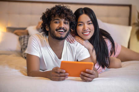 Man with tablet and hugging woman lying on bed