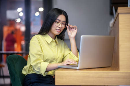 Cute asian young woman in eyeglasses at the laptop having coffeeの写真素材