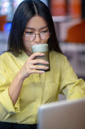 Cute asian young woman in eyeglasses at the laptop having coffeeの写真素材