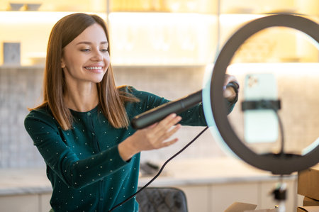 Smiling young hairdresser holding hair straighntener and looking contented