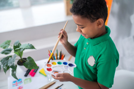 Enjoyment. Smiling dark-skinned boy of primary school age in green tshirt looking at palette with paints and brush in hands standing near table in bright roomの写真素材