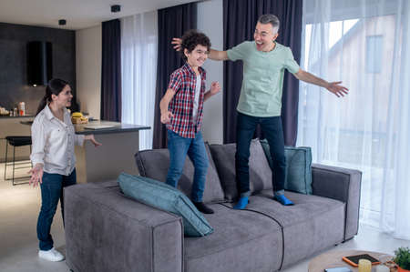 Dad and son standing on couch and woman watchingの写真素材