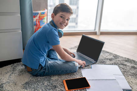Cheerful pupil sitting on the carpet during his online lesson