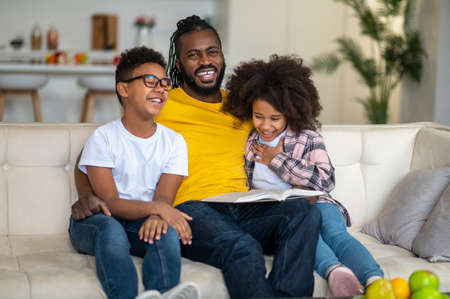 Dad with book hugging children laughing sitting on sofaの写真素材