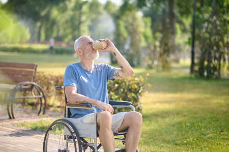 A man in a wheel chair sitting in the park and having coffeeの写真素材