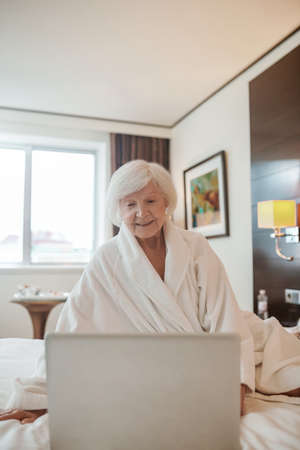 An old woman with a laptop sitting on a bed in a hotel roomの写真素材