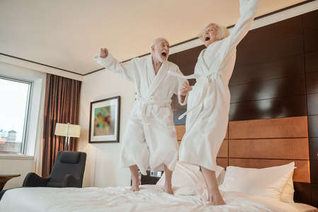 Senior couple in white robes jumping on bed and looking happyの写真素材
